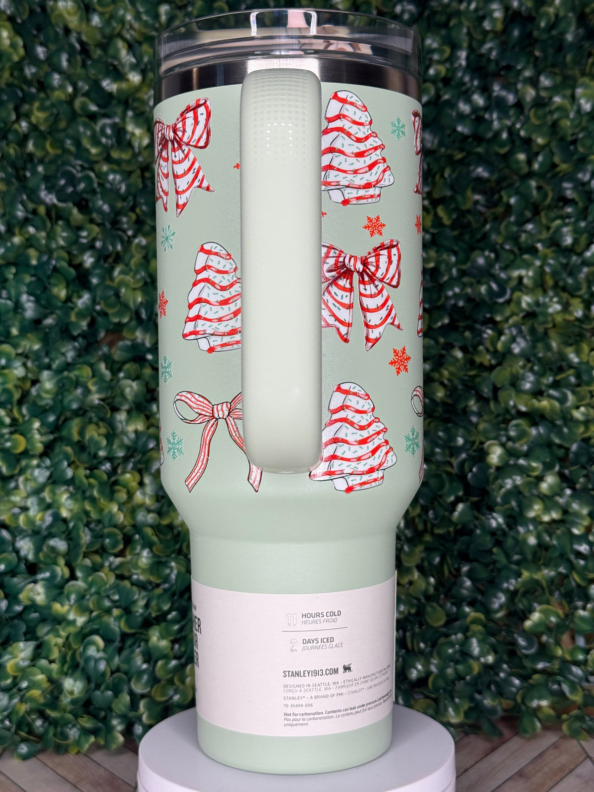 A green insulated tumbler with a festive pattern of red and white bows and snowflakes on it, placed on a surface with green foliage.